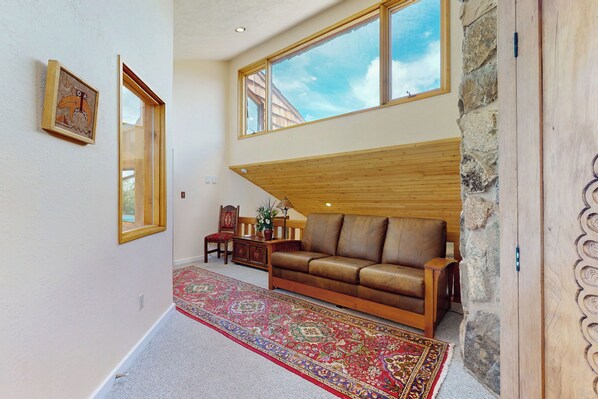 A spacious living room with a high ceiling, a large window, a brown leather sofa, a patterned rug, and a wooden ceiling beam. - 4 BR Home w/ Pool, Hot Tub, Fireplaces, Grills (Arroyo Seco)