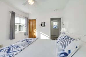 Et hotelværelse med seng, ventilator, et vindue med gardiner, en dør og et spejl på væggen.