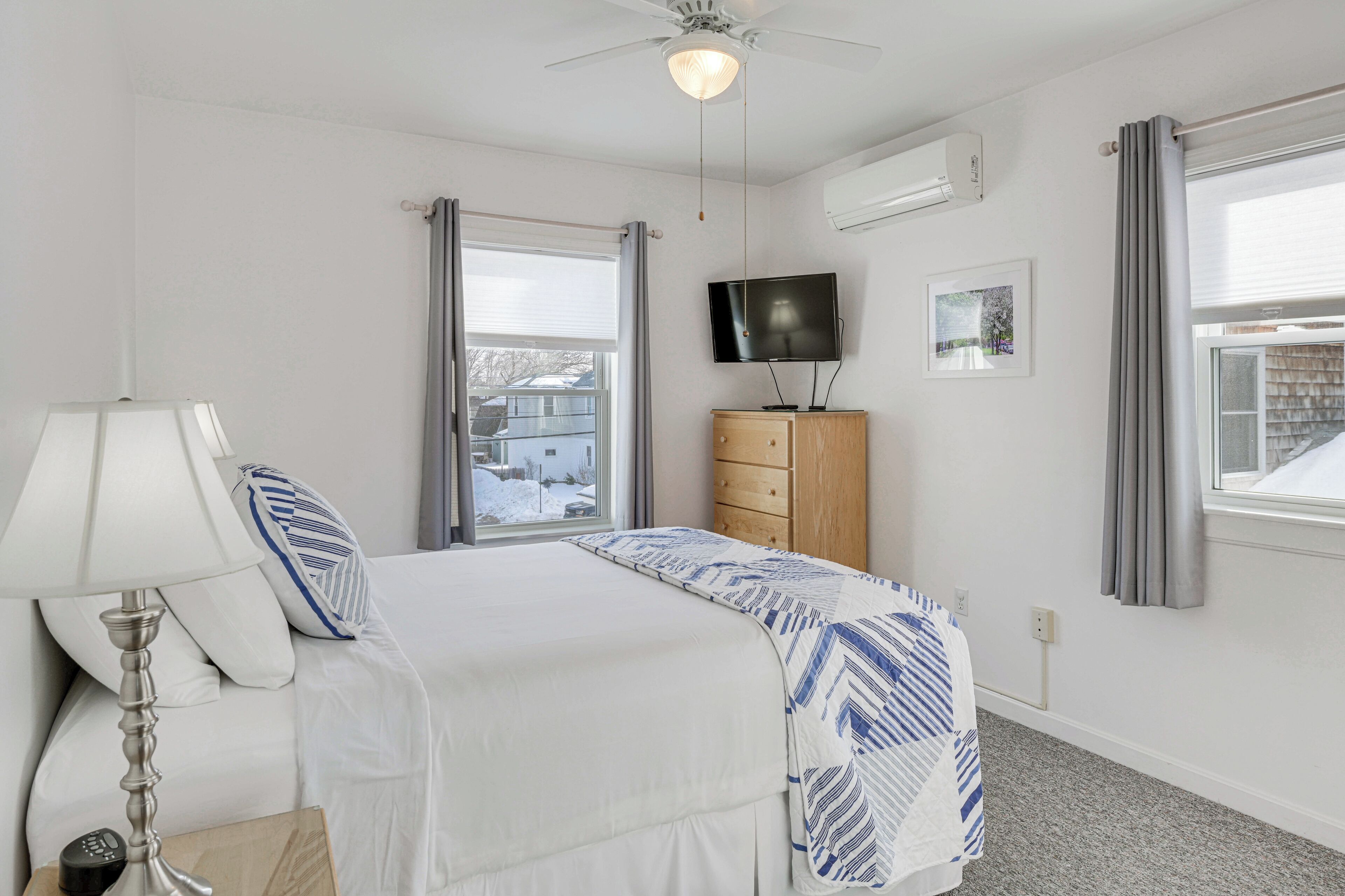 Un dormitorio con cama, mesita de noche con lámpara, cómoda, televisor y ventana con cortinas.