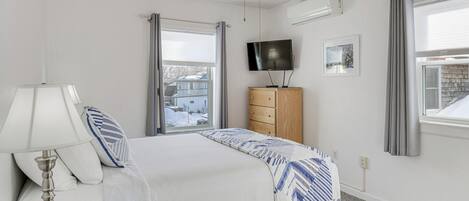Un dormitorio con cama, mesita de noche con lámpara, cómoda, televisor y ventana con cortinas.