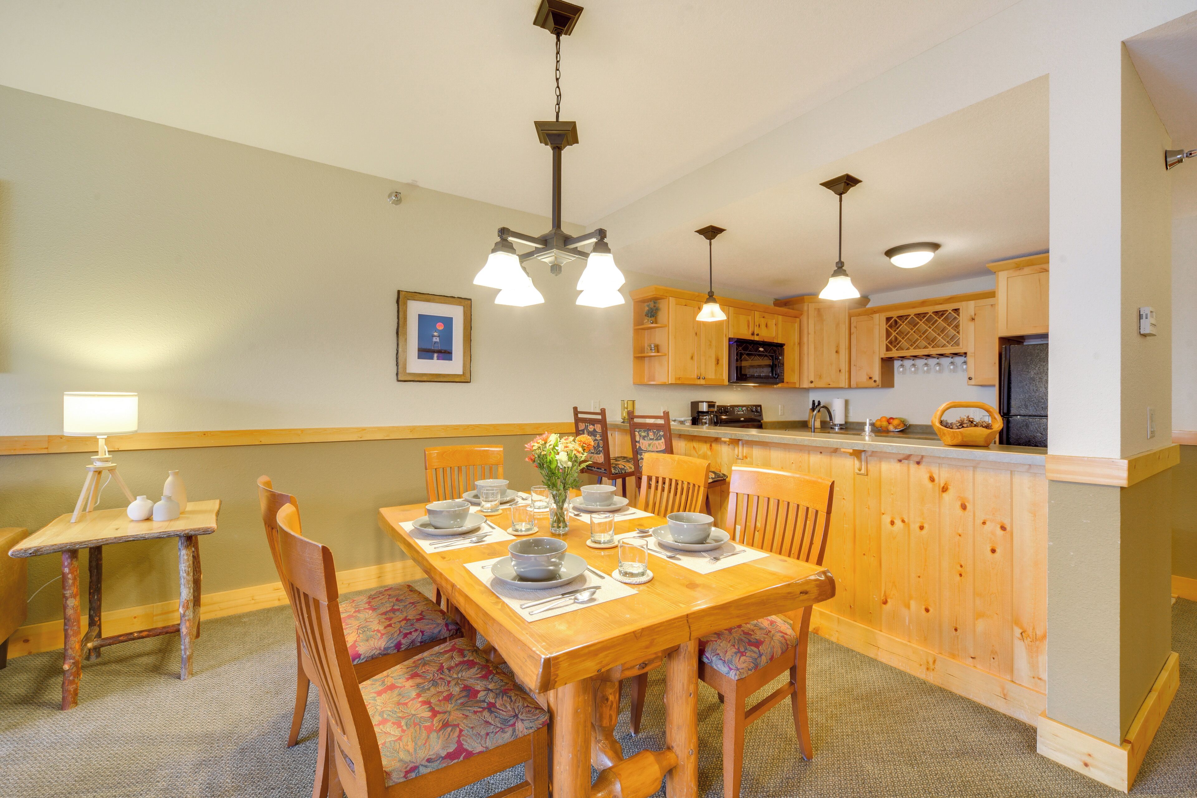 Un comedor con una mesa de madera dispuesta para cuatro personas, rodeada de sillas con tapicería floral. Se ve una cocina con armarios de madera y una barra en el fondo.