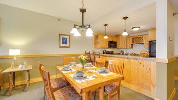Un comedor con una mesa de madera dispuesta para cuatro personas, rodeada de sillas con tapicería floral. Se ve una cocina con armarios de madera y una barra en el fondo.