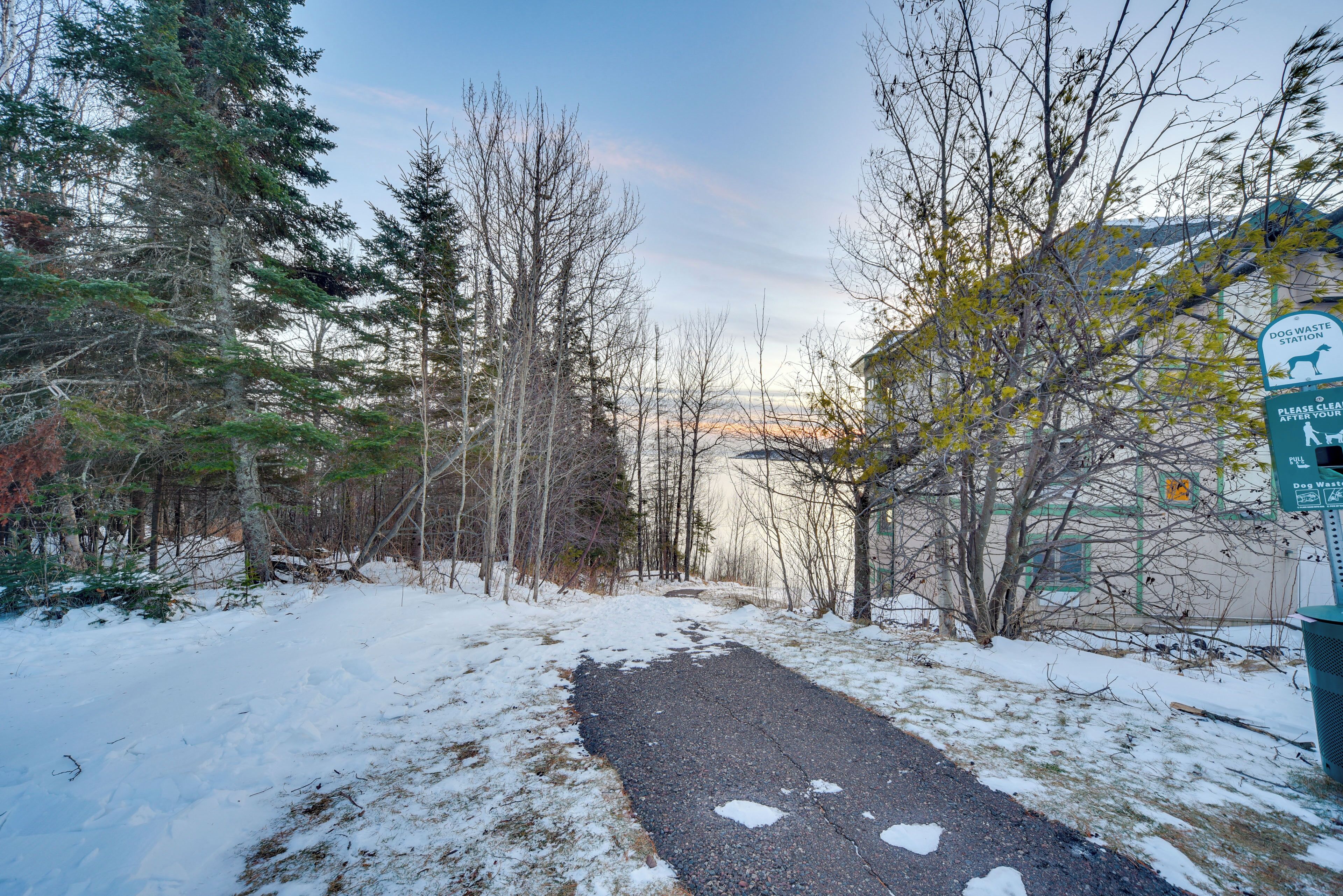 A snow-covered path leading through a forested area with a dog waste station sign.
