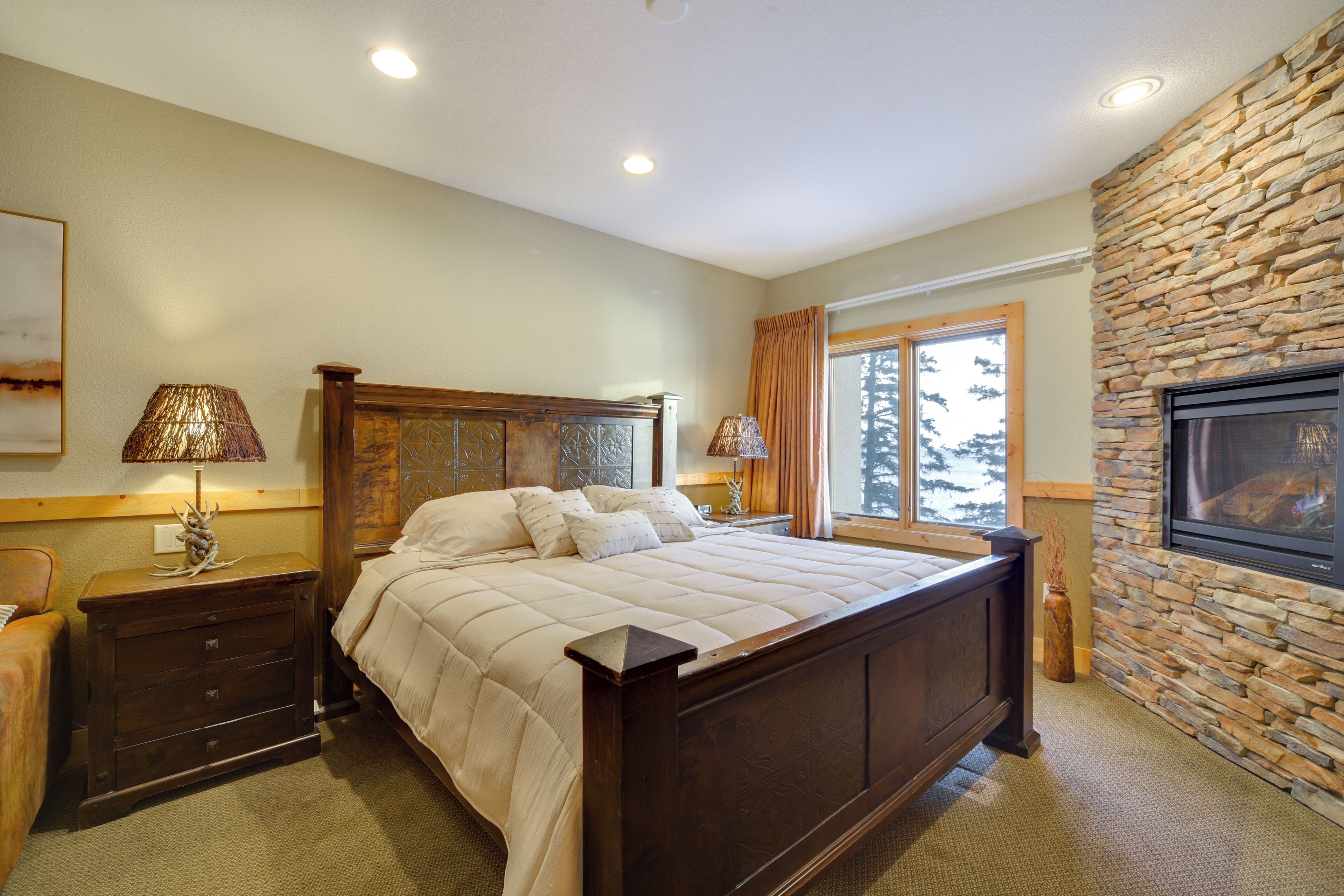A bedroom with a large bed, wooden headboard, two bedside tables with lamps, a stone fireplace, and a window with curtains.