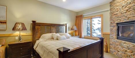 A bedroom with a large bed, wooden headboard, two bedside tables with lamps, a stone fireplace, and a window with curtains.