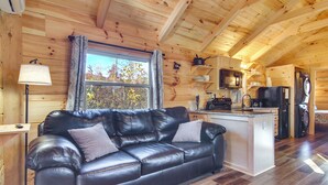 Interior acogedor de una cabaña con sofá de cuero, paredes de madera, una zona de cocina y una ventana con vistas a los árboles.