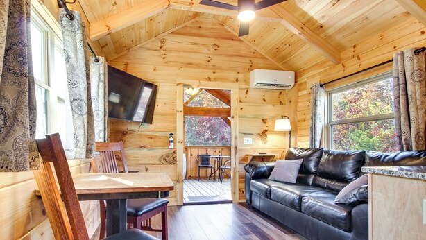 Interior de una cabaña de madera con una mesa de comedor de madera, sillas, un sofá de cuero y un televisor montado en la pared.