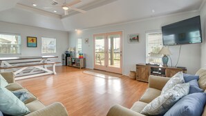 A spacious living room with hardwood floors, a ceiling fan, a large TV, and a sliding glass door leading to a balcony. - Steps to Sea! Crystal Beach Home w/ Furnished Deck (Port Bolivar)