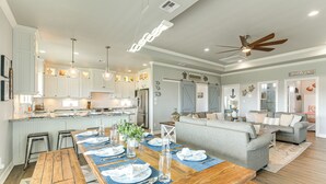 Un vaste espace de vie comprenant une grande table à manger en bois, disposée pour accueillir quatre personnes, un canapé gris et un ventilateur de plafond.