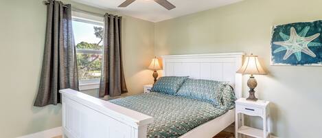 A bedroom with a bed, a ceiling fan, a window with curtains, a nightstand with a lamp, and a wall with a starfish decoration.