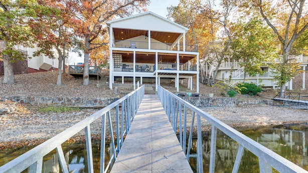 A wooden deck leading to a two-story building with a balcony, surrounded by trees and a body of water. - Waterfront Family Getaway in Lake Ozark w/ Dock (Lake Ozark)