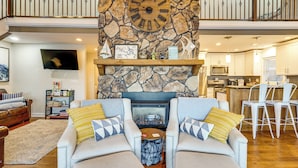 A spacious living room with a stone fireplace, comfortable seating, a flat-screen TV, and a kitchen in the background. - Waterfront Family Getaway in Lake Ozark w/ Dock (Lake Ozark)