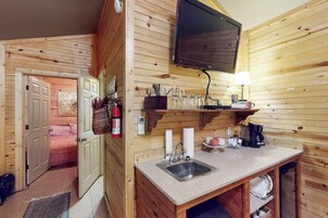 Interior de una cabaña de madera con zona de cocina que incluye un lavabo, un televisor montado en la pared y una estantería con cristalería.