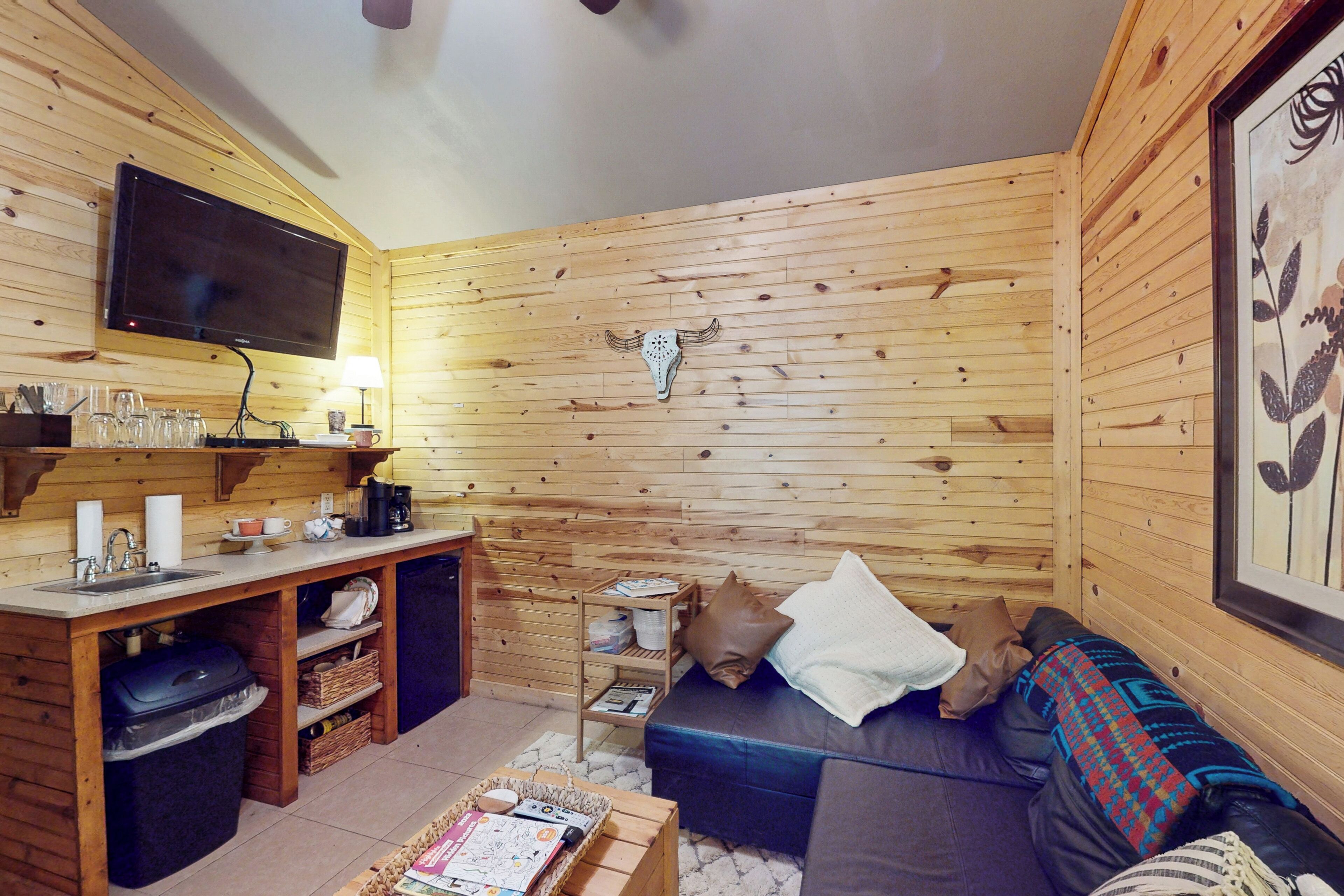 A wooden cabin interior with a TV mounted on the wall, a sofa, a coffee table, and a shelf with various items.