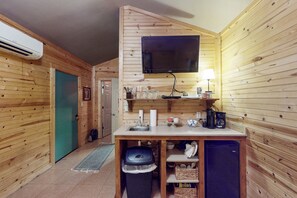Eine kleine Küche mit Holzwänden, einem an der Wand montierten Fernseher und grundlegender Ausstattung, darunter ein Spülbecken, eine Kaffeemaschine und Ablageflächen.