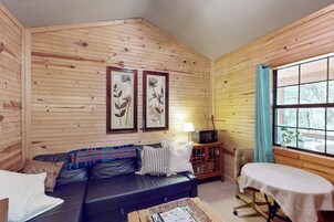 Un interior de cabaña acogedor con una pared de madera, un sofá de cuero, una pequeña mesa y una ventana con cortinas.