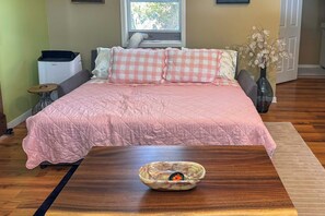 Ein Schlafzimmer mit Bett, Nachttisch, Fenster, Heizung, einer Blumenvase und Holzboden.