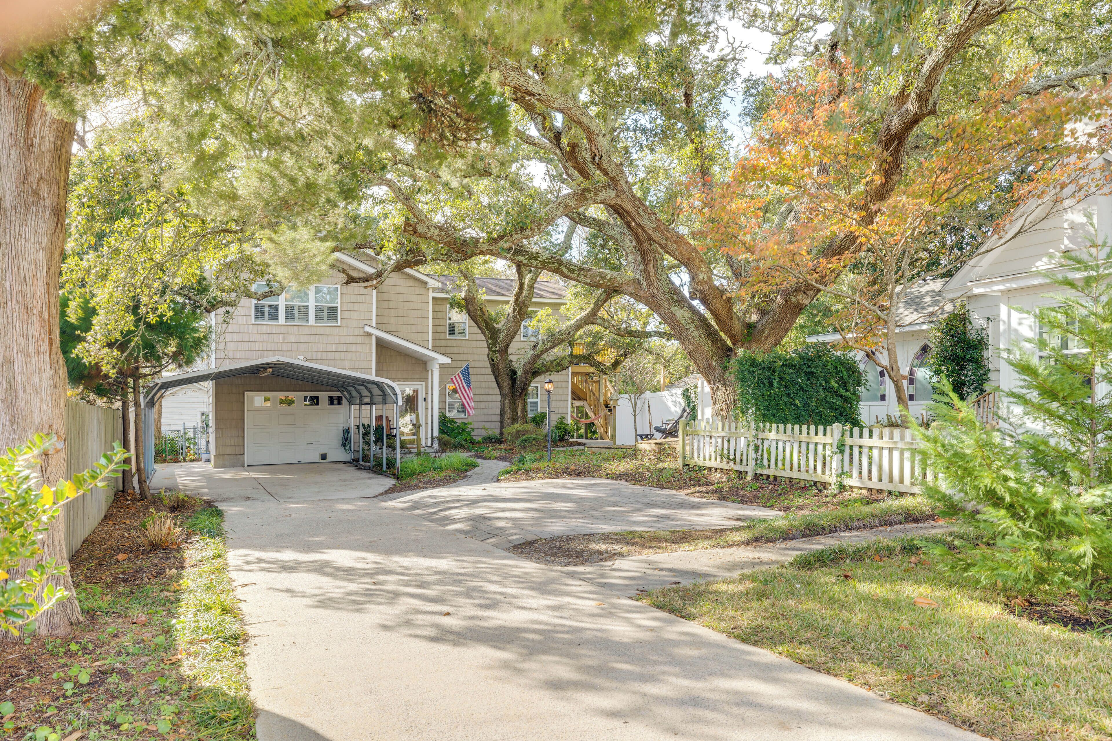 A residential driveway leading to a garage with a covered carport, surrounded by mature trees and a white picket fence.