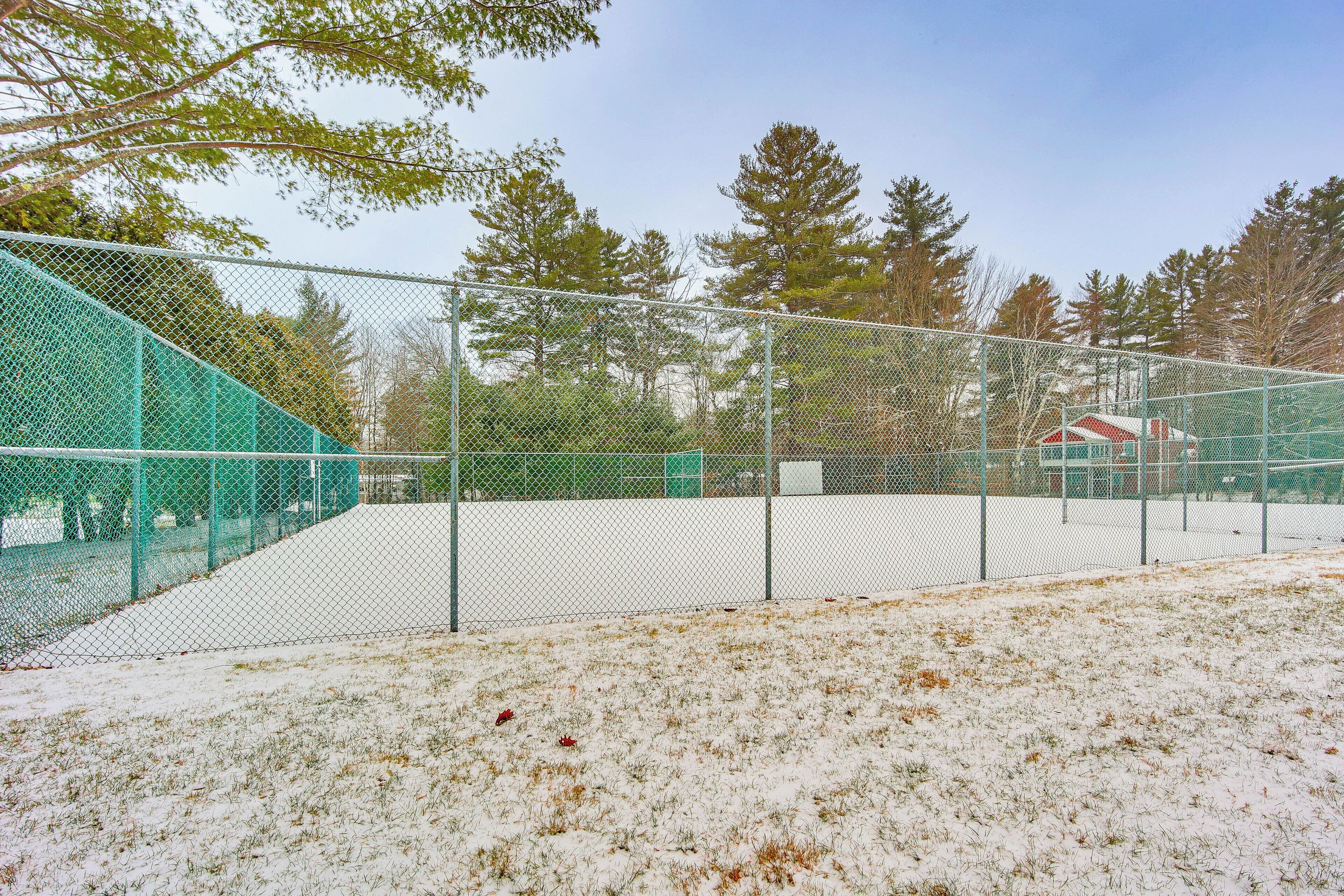 Cancha de tenis rodeada por una valla, con nieve en el suelo y árboles al fondo.