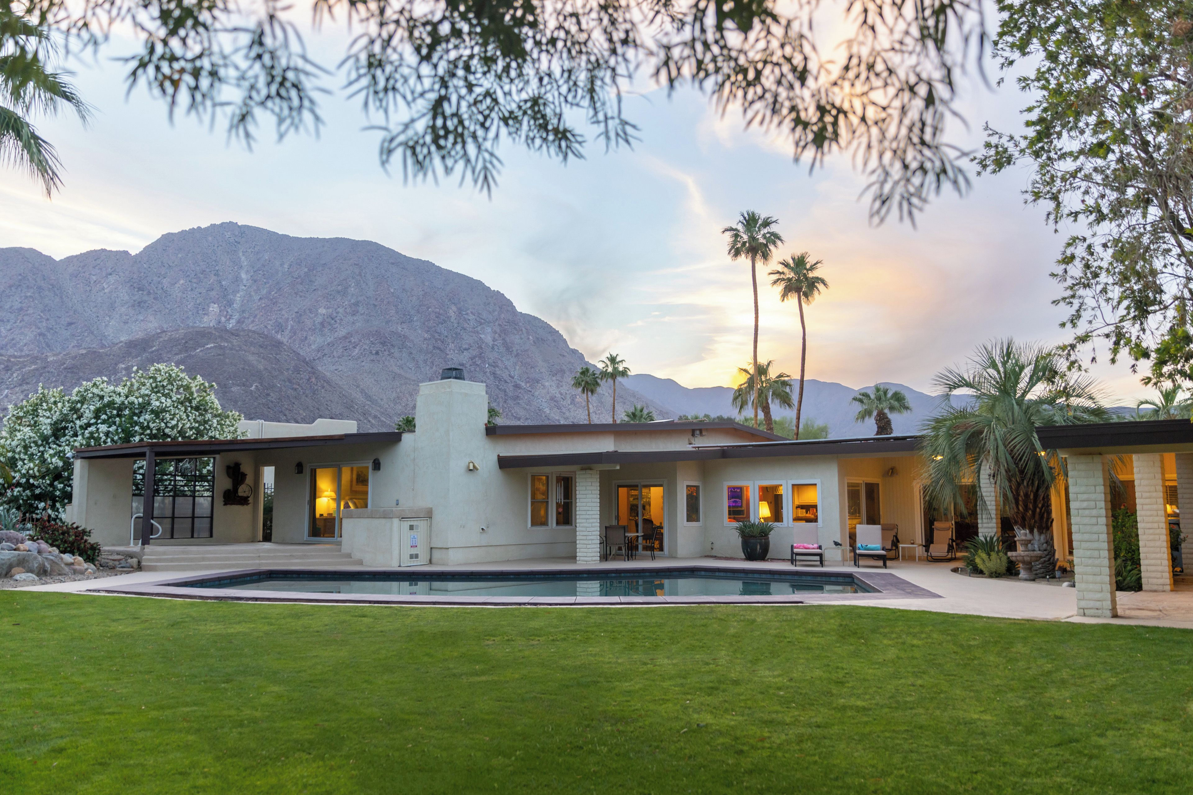 Una casa moderna con piscina, rodeada de césped cuidado y palmeras, con montañas al fondo.