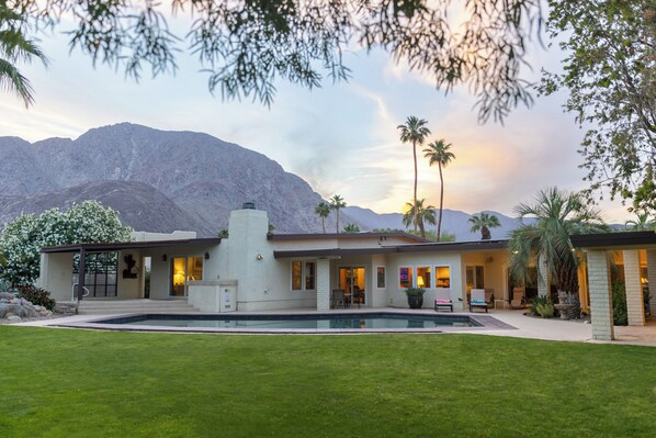 Une maison moderne avec une piscine, entourée d’une pelouse bien entretenue et de palmiers, avec des montagnes en toile de fond.