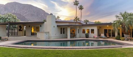 A modern house with a swimming pool, palm trees, and mountains in the background.