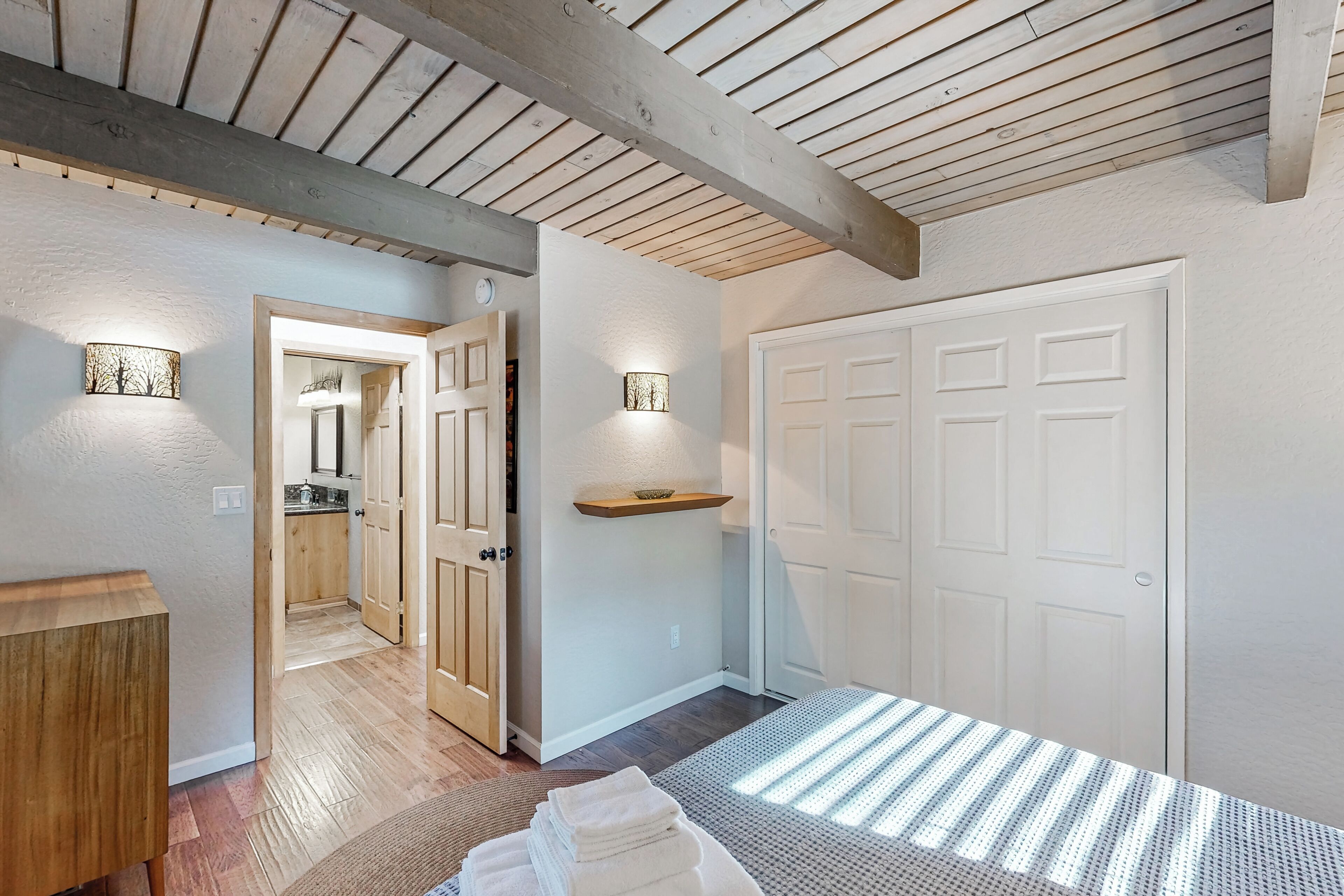 A bedroom with a wooden ceiling, a bed with white linens, a nightstand, a shelf with a bowl, and a door leading to a bathroom.
