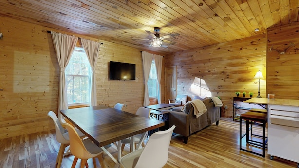 Een gezellig interieur van een chalet, met een houten plafond, een eettafel met stoelen, een bank, een tv aan de wand en een klein keukentje.
