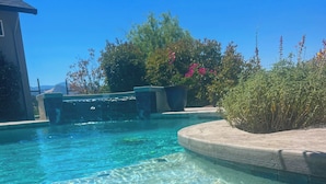 Una piscina cristalina con un borde de piedra, rodeada de follaje y una casa al fondo.