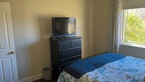 Un dormitorio con cama, cómoda, televisor encima de la cómoda, ventana con cortinas y puerta.