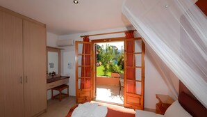 A bedroom with a bed, a desk, a mirror, and a view of a garden through an open door.