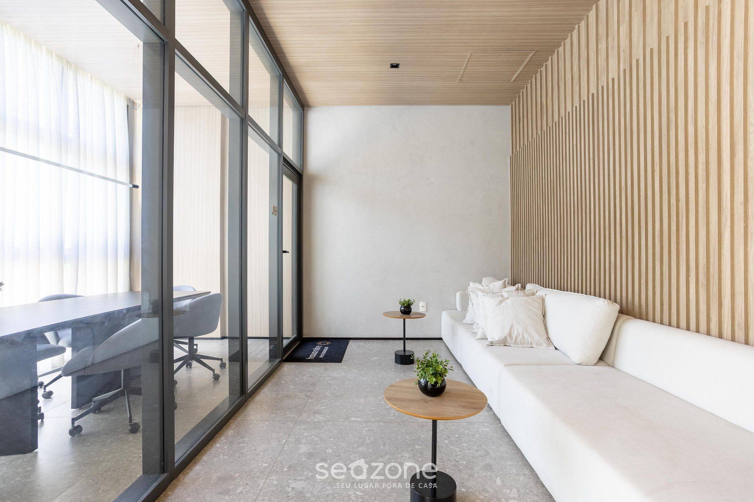 A modern office space with a glass partition, a conference table, a sofa, and two small tables with plants.