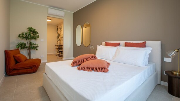 A bedroom with a large bed, two pillows, a bedside table with a lamp, a chair, a plant, and a mirror on the wall. - Daplace - Ettore Apartment (Milano)