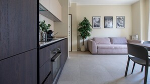 Une cuisine moderne avec des meubles de couleur sombre, un four encastré et une machine à café. Le salon comprend un canapé beige et une table à manger avec des chaises.