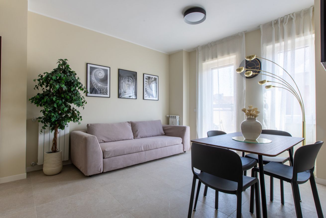 A modern living room with a beige sofa, a dining table with chairs, a potted plant, and framed artwork on the wall.