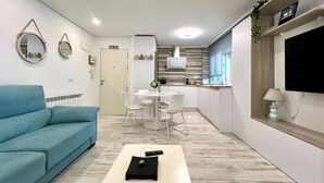 A modern living room with a blue sofa, a coffee table, and a TV on the wall. There is a kitchen area with white cabinets and a dining table with chairs. - 3 rooms apartment close beach (Santander)