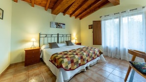 Un dormitorio con techo de madera, una cama con colcha, dos mesitas de noche con lámparas, una silla y una ventana con cortinas.