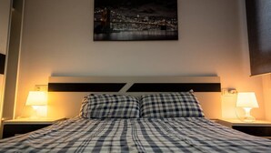 Una camera d'albergo con un letto, due abat-jour, una coperta a quadretti e un dipinto con una vista sulla città.