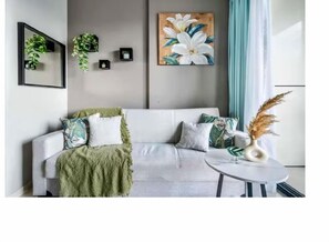 Ein modernes Wohnzimmer mit einem grauen Sofa, einer grünen Tagesdecke und einer weißen Vase mit Pampasgras.
