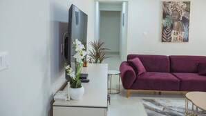 A modern living room with a purple sofa, a flat-screen TV, a white coffee table, and a painting on the wall. - The Oasis Airport Pick up Wifi Central (Accra)