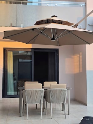 En modern utomhusplats för måltider med ett bord och stolar, ett stort parasoll och utsikt över staden genom en glasdörr.