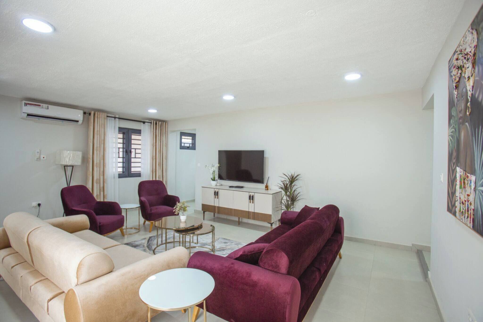 A modern living room with a beige sofa, a red sofa, two purple armchairs, a small white table, a TV on a white cabinet, and a window with curtains.