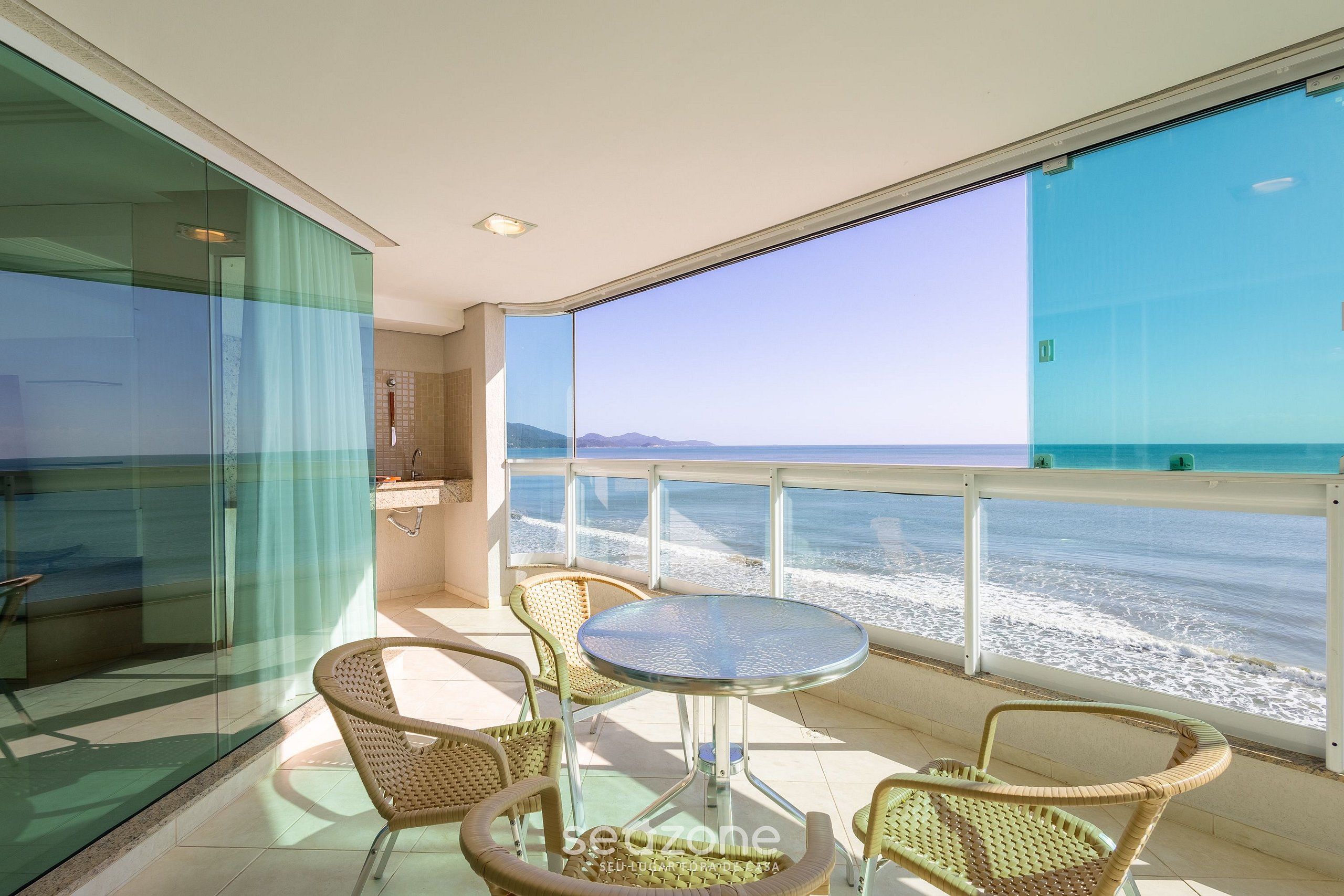 Ein Balkon mit Korbstühlen und einem Glastisch, mit Blick auf einen Strand und Wellen.