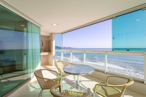 Un balcon avec des fauteuils en osier et une table en verre, donnant sur une plage où l’on peut admirer les vagues.