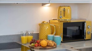 A kitchen with a yellow toaster, a black microwave, and a coffee maker. There are two glasses of orange juice, a plate with strawberries, grapes, and an orange, and two blue cups. - Bright Stylish Studio Apartment (Stretford)