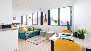 A modern apartment with a living area, kitchen, and dining space. It features large windows, a guitar, and a plant. - Bright Stylish Studio Apartment (Stretford)