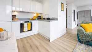 A modern kitchen with white cabinets, a microwave, and a kettle. There is a wooden floor and a green sofa with a yellow pillow in the living area. - Bright Stylish Studio Apartment (Stretford)