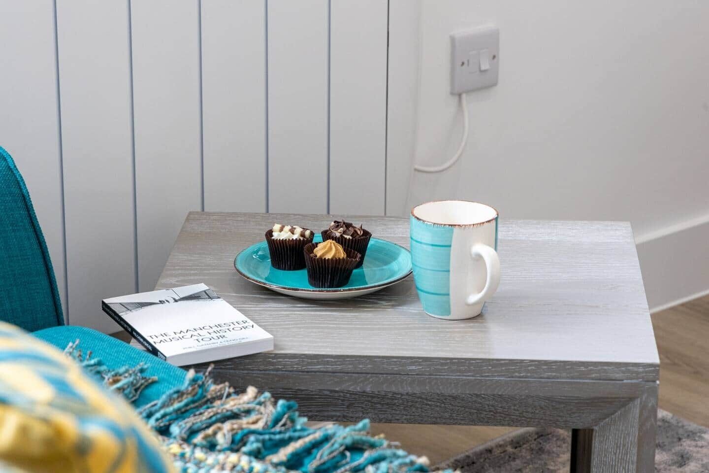 Een klein tafeltje met een bord cupcakes en een mok, een boek getiteld 'The Manchester Musical History Tour', en een stopcontact op de muur.