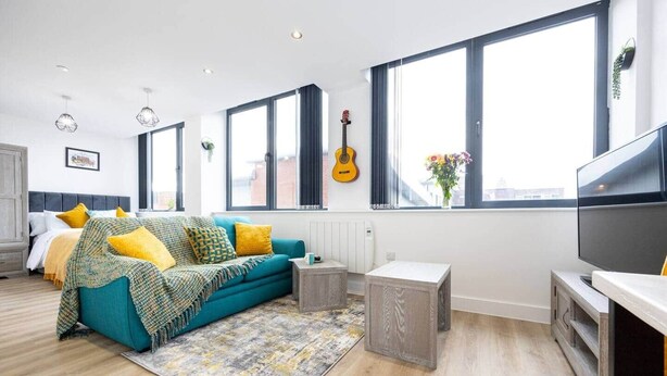 A modern living room with a teal sofa, yellow cushions, a grey rug, and a guitar on the wall. - Bright Stylish Studio Apartment (Stretford)