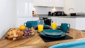 A breakfast setup with croissants, grapes, and orange juice on a wooden table in a modern kitchen with white cabinets and a black coffee machine. - Luxurious Stylish Apartment (Stretford)